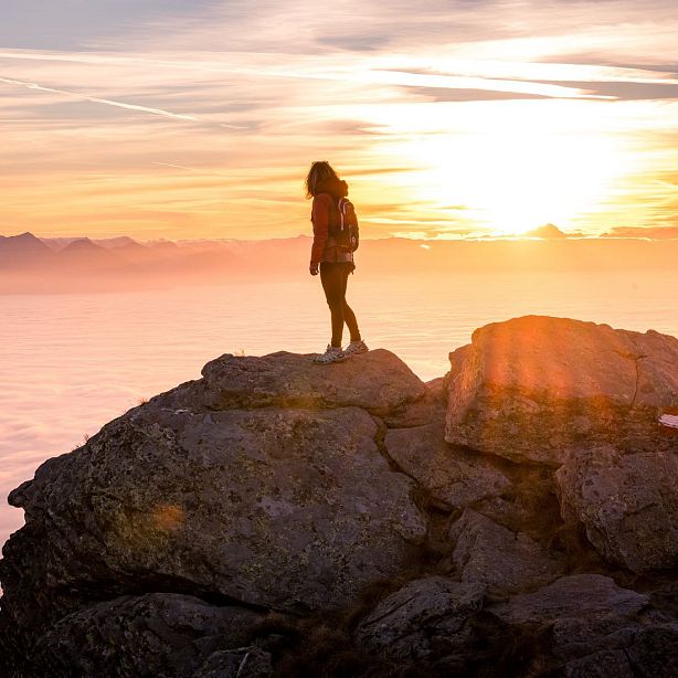 Person standing on rocky cliff at sunrise, overlooking clouds and distant mountains, creating a serene and majestic landscape.
