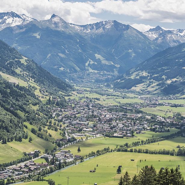 Picturesque alpine village in a green valley, bordered by forested mountains and distant snow-capped peaks under a clear sky.
