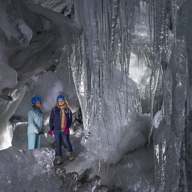 Zwei Personen in blauer Winterkleidung stehen in einer Eishöhle mit riesigen Eiszapfen. Sie tragen Helme und sehen beeindruckt aus. Die Höhle ist frostig und hell.