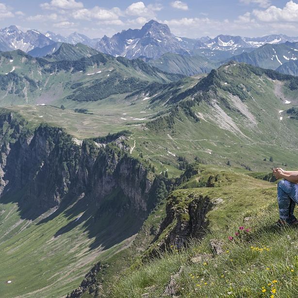 Majestic mountains with green valleys and rocky cliffs under clear skies. A person sits on the edge, taking in the scenic view.