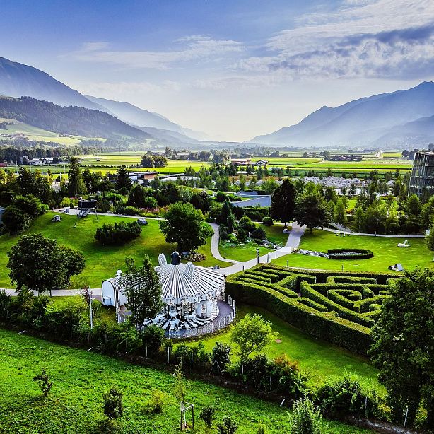 Scenic view of green fields, a small carousel, and a hedge maze with mountains in the background under a clear blue sky.