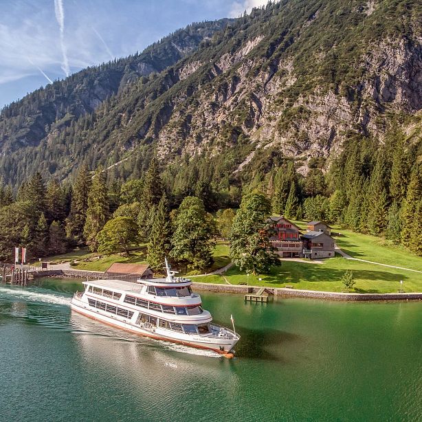 A boat cruises on a clear lake, surrounded by towering mountains and lush trees, with wooden houses nearby, under a blue sky.