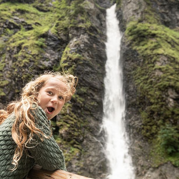 Child with surprised expression in front of a tall waterfall cascading down a green, rocky cliff.
