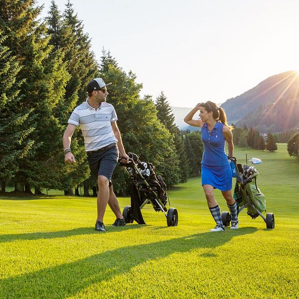 A man and woman walk on a sunny golf course with their golf bags, surrounded by trees and mountains.