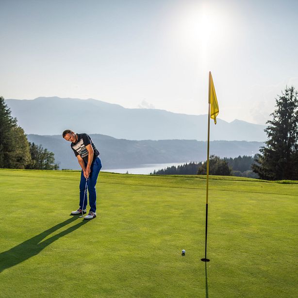 A golfer putts on a lush green course with a yellow flag, surrounded by trees and mountains in the background under a clear sky.