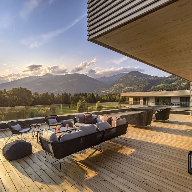 A spacious terrace with modern furniture and a panoramic view of mountains at sunset, bathed in warm light.