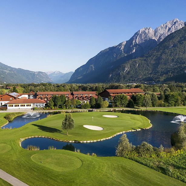 A golf course with green grass, lakes, and fountains set against a backdrop of mountains and a rustic building.