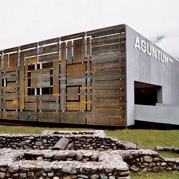 Modern museum building with rusty, industrial facade, displaying 'Aguntum'. Beneath are ancient stone ruins. The architecture contrasts with historic remains.