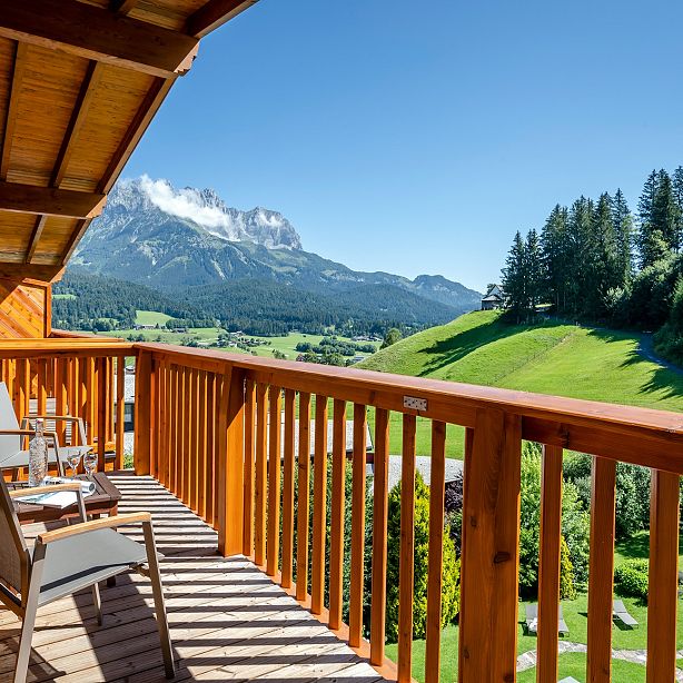 Balkon mit Holzgeländer und Stühlen, Blick auf grüne Wiesen und Wälder, dahinter hohe Berge mit schneebedeckten Gipfeln unter einem klaren blauen Himmel.