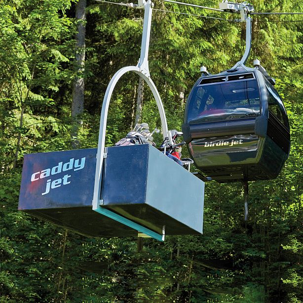 A gondola lift carries golf gear in a wooded area, featuring a Caddy Jet container. Bright daylight highlights the lush green surroundings.