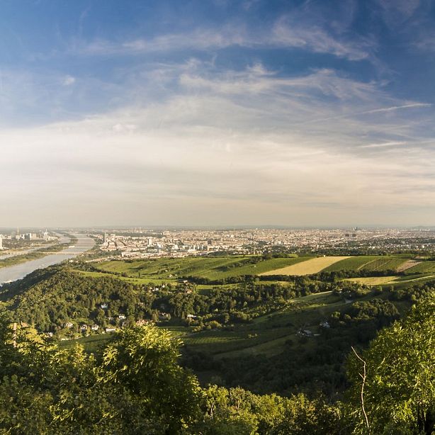 Panoramic view of a city by a river, surrounded by green hills and under a partly cloudy sky, showcasing the beauty of the natural and urban landscapes.