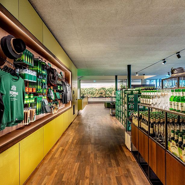 Ein stilvoller Getränkeladen mit hölzernem Boden, flankiert von Regalen voller grüner Merchandise-Artikel auf der linken Seite und Getränkekästen rechts.