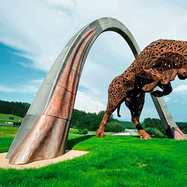 Large metal sculpture of a leaping animal under a modern arch on a grassy field, with trees and clear blue sky in the background.