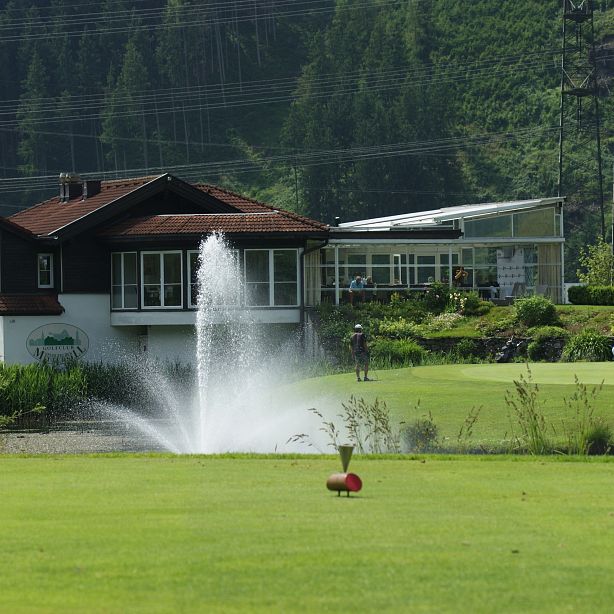 Golfszene mit saftigem Grün, einem zentralen Brunnen, einem stilvollen Clubhaus im Hintergrund und einem bedeckten Himmel. Im Vordergrund ein Golfschläger auf dem Rasen.