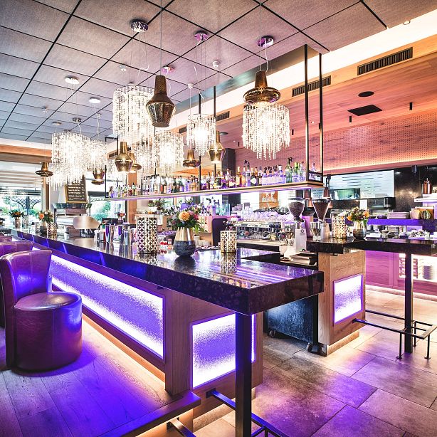 Elegante Bar mit modernem Design, beleuchtet durch warme Lichter und Kristallleuchten. Stilvolle Barhocker und ein gut bestücktes Regal mit Flaschen im Hintergrund.