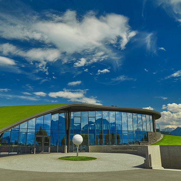 Modernes Gebäude mit begrüntem Dach und Glasfassade vor blauem Himmel mit Wolken, umgeben von Bergen in einer landschaftlich reizvollen Umgebung.
