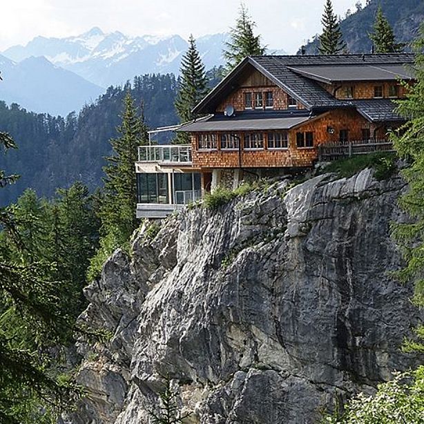 A wooden house sits atop a rocky cliff surrounded by pine trees, with mountains in the background. It has large windows and offers a scenic, secluded view.