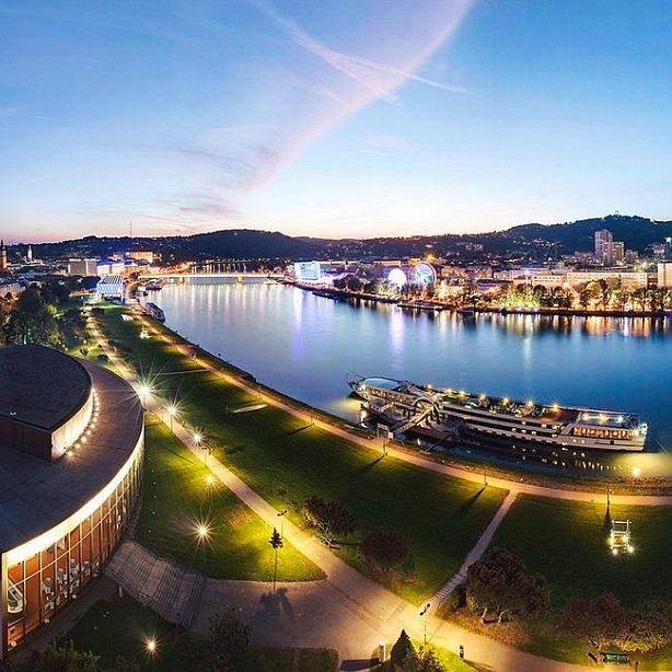 A scenic view of a river with illuminated city buildings and a boat at dusk, surrounded by lights and hills, reflecting a peaceful urban landscape.