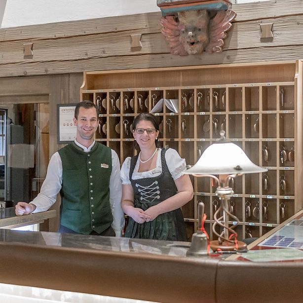 Two staff in traditional Bavarian attire at a hotel reception desk, with a key rack in the background, creating a welcoming atmosphere.