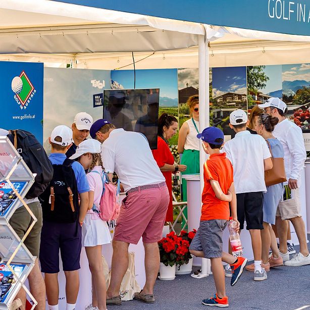 Gruppe von Kindern und Erwachsenen bei einer Messe, umgeben von Informationsständen. Sie schauen auf einen Bildschirm und tragen sommerliche Kleidung und Caps.