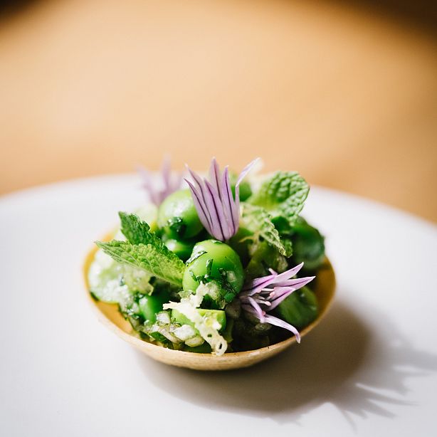 Eine kleine, dekorative Schüssel mit einem frischen grünen Salat aus Erbsen, frischen Minzblättern und lilafarbenen essbaren Blüten, auf einem hellen Hintergrund.