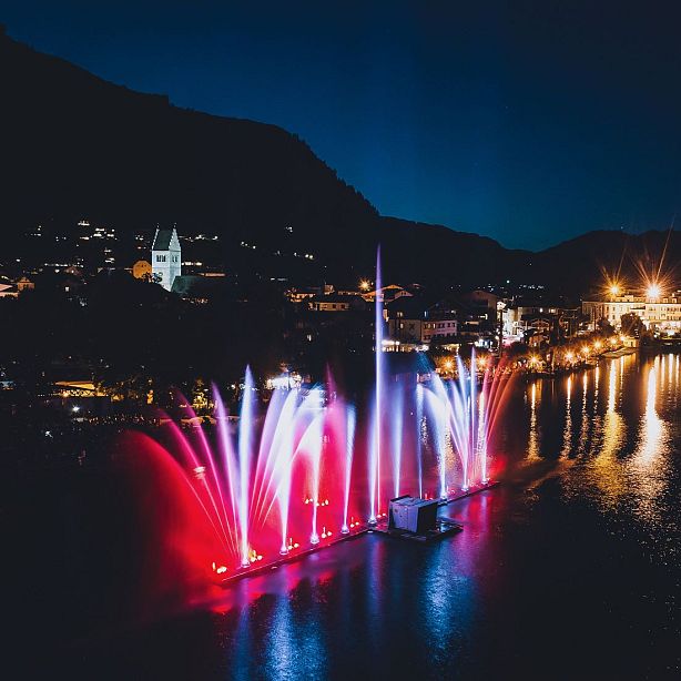 Nighttime fountain show with colorful lights over a lake, surrounded by town buildings and hills. Illuminated water jets in red, blue, and purple hues.