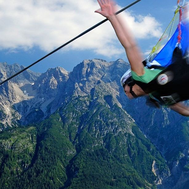 Person zip-lining across a mountain range with rugged peaks in the background and a clear blue sky above. The scene captures adventure and adrenaline.