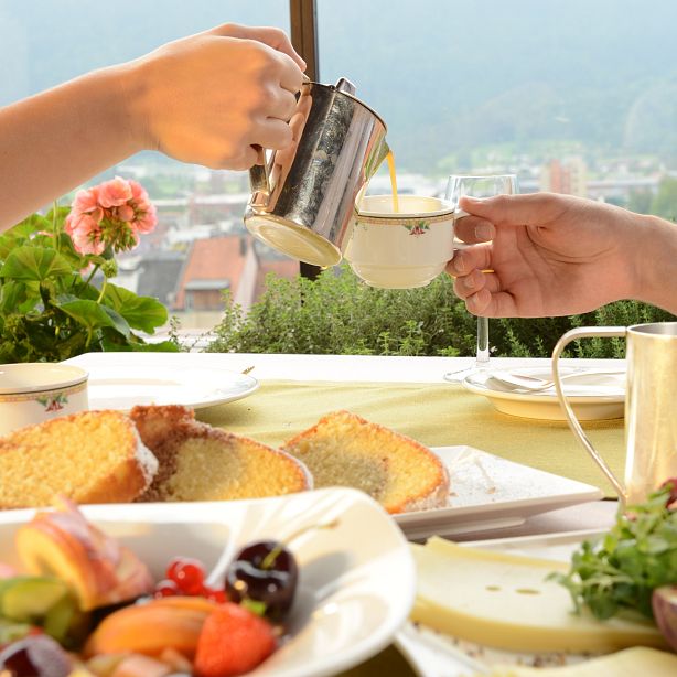 Breakfast scene on a terrace with mountains; a hand pouring coffee, with dishes of cake, cheese, fruits. Greenery and pink flowers in the background.