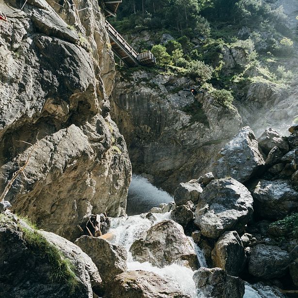 A waterfall cascades through rocky mountains, surrounded by greenery and a wooden bridge. Sunlight filters through, highlighting the natural beauty and serenity.