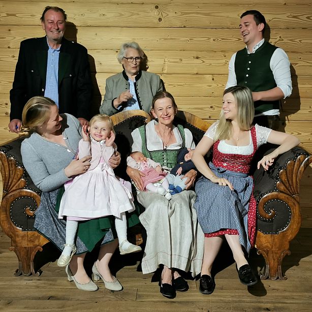 Eine fröhliche Familie in traditioneller bayerischer Tracht sitzt in einem Raum mit Holzwänden. Drei Generationen sind auf einem Sofa versammelt.
