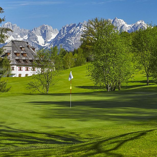 Golf course with a flagstick, lush green grass, trees, and a mountain range in the background with a chalet visible to the left.