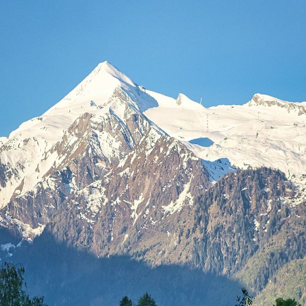 A snow-capped mountain peak with rugged slopes and patches of snow, set against a clear blue sky, creating a picturesque natural scene.
