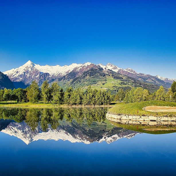 Snow-capped mountains with green slopes are reflected in a calm lake, surrounded by trees, under a blue sky.