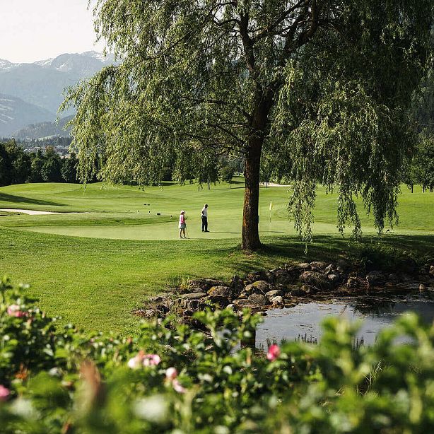 Zwei Personen spielen Golf auf einem gepflegten Golfplatz mit grünen Fairways und einem großen Baum. Im Vordergrund ist ein Teich zu sehen.