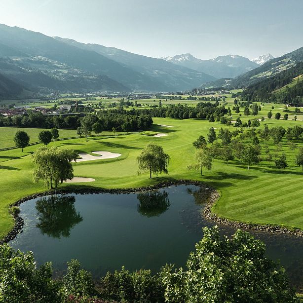 Grüner Golfplatz in einer alpinen Landschaft mit Bergen im Hintergrund. Der Platz hat einen Teich, Sandbunker und gepflegte Rasenflächen unter blauem Himmel.