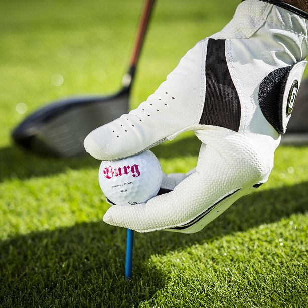 Ein Golfspieler platziert einen Golfball auf einem blauen Tee auf dem Rasen. Der Spieler trägt einen weißen Handschuh. Ein Golfschläger ist im Hintergrund sichtbar.