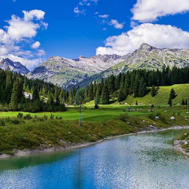 Ein klarer Bergsee spiegelt den blauen Himmel und die weißen Wolken wider, umgeben von grünen Wäldern und majestätischen Bergen in der Ferne.