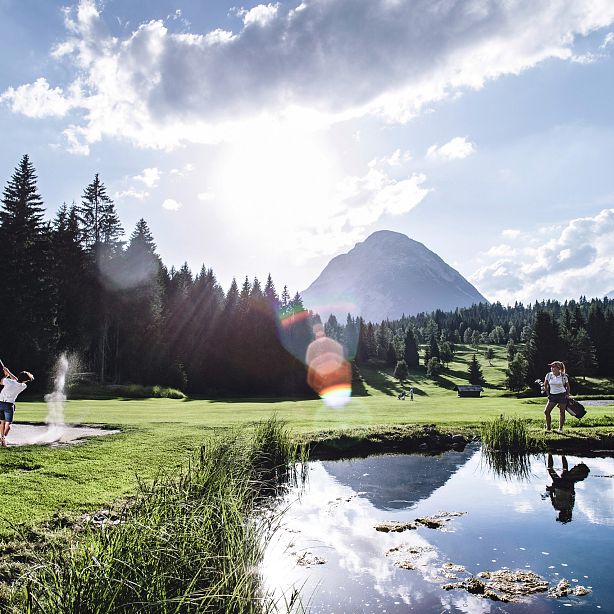 A golfer swings as two horseback riders pass by, set against a mountain backdrop with sunlight breaking through clouds, reflecting in a nearby pond.