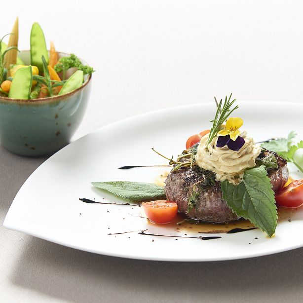 A gourmet dish on a white plate with artistic presentation, surrounded by garnish and herbs. A bowl of fresh salad with green veggies in the background.