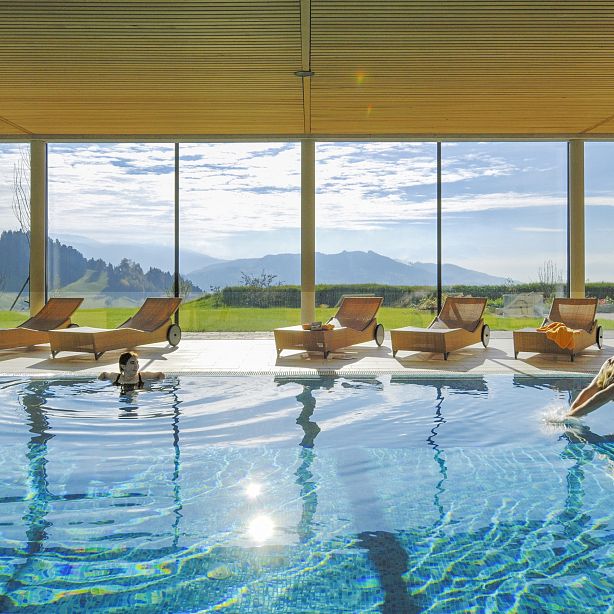 Ein moderner Innenpool mit großen Fenstern und Blick auf eine hügelige Landschaft. Mehrere Liegestühle stehen am Beckenrand, während eine Person ins Wasser springt.