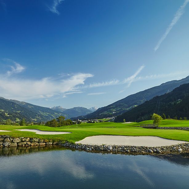 Golf course in a mountain valley with sand bunkers and a small pond, surrounded by lush greenery and mountains under a clear blue sky.