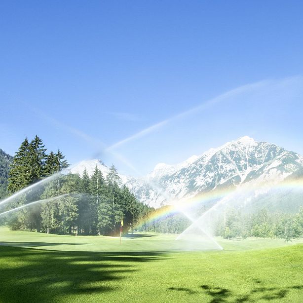 Green field with water sprinklers creating arcs and a rainbow, set against a backdrop of pine trees and snow-capped mountains under a clear blue sky.