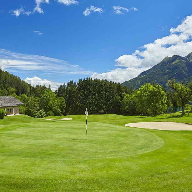 A scenic golf course with a green field, sand bunker, and flag, surrounded by mountains and trees under a blue sky with white clouds.