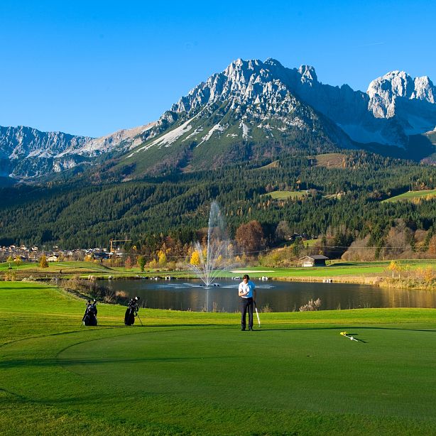 Scenic golf course with golfers on a green, surrounded by majestic mountains and a blue sky, creating a tranquil and beautiful outdoor setting.