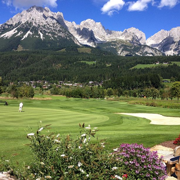 Ein gepflegter Golfplatz mit Spielern im Vordergrund. Im Hintergrund erheben sich imposante, schneebedeckte Berge unter einem klaren, blauen Himmel.