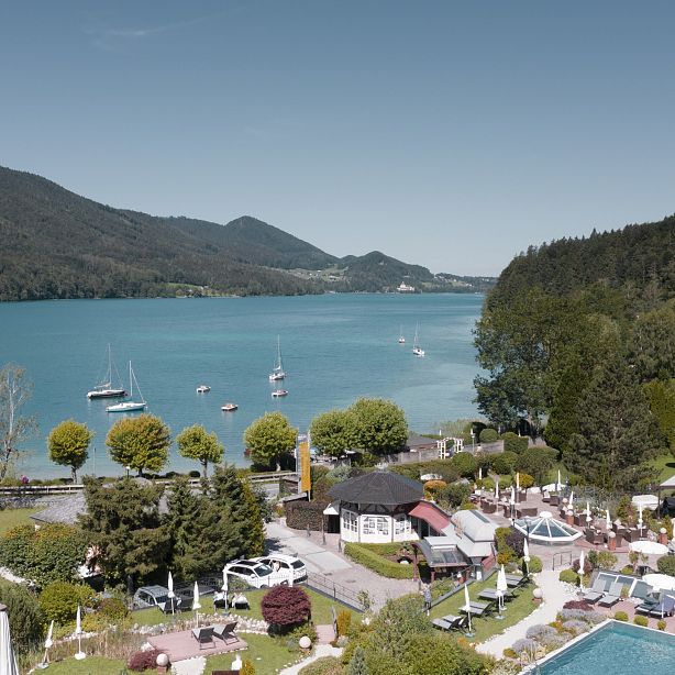 Tranquil lake with boats and surrounding hills. Foreground features pool and lounging area amid greenery, under clear blue sky.