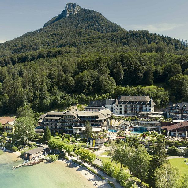 Ein malerischer See mit einem Hotel am Ufer, umgeben von üppigen Wäldern und einem dominanten Berg im Hintergrund unter einem klaren blauen Himmel.