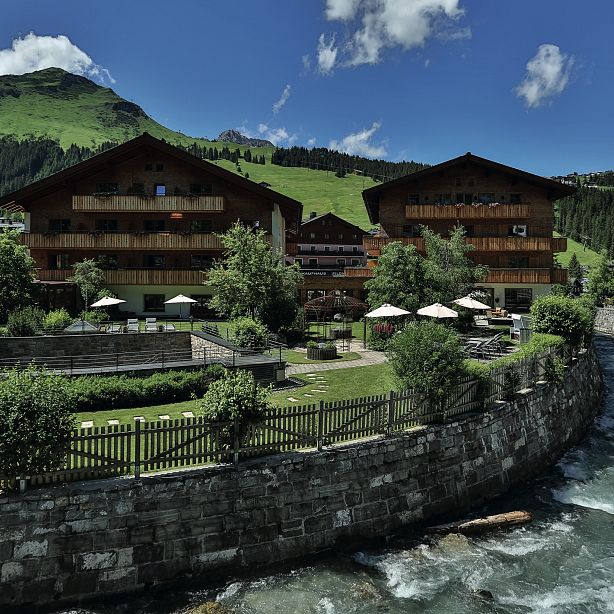 Mountain resort buildings by a river, surrounded by greenery and mountains under a blue sky with clouds.