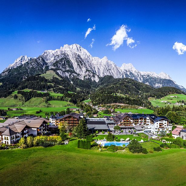 A scenic view of a mountain resort surrounded by green hills and tall mountains under a clear blue sky.
