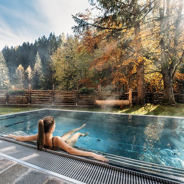 A person relaxes in a steaming outdoor pool amidst an autumnal forest with colorful leaves, under a clear blue sky.